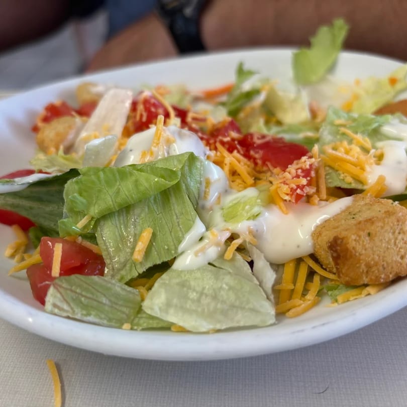 A diner side salad with lettuce, tomatoes, shredded cheese, croutons, and ranch dressing.